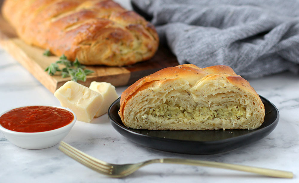 Whole Four Cheese and Herb pastry on serving platter with slice of pastry on plate next to it. Pieces of cheese and sprigs of herbs next to it as garnish with a bowl of marinara sauce.