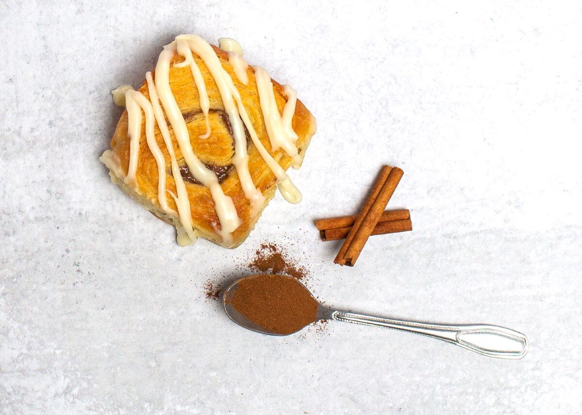 Cinnamon pastry roll next to 2 cinnamon sticks and a spoon with ground cinnamon