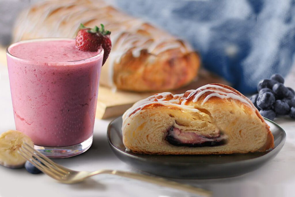 Whole Blueberry & Cream Cheese pastry on cutting board. Slice of pastry on a plate with blueberries next to it as garnish. Glass with Strawberry Banana Smoothie to the side.
