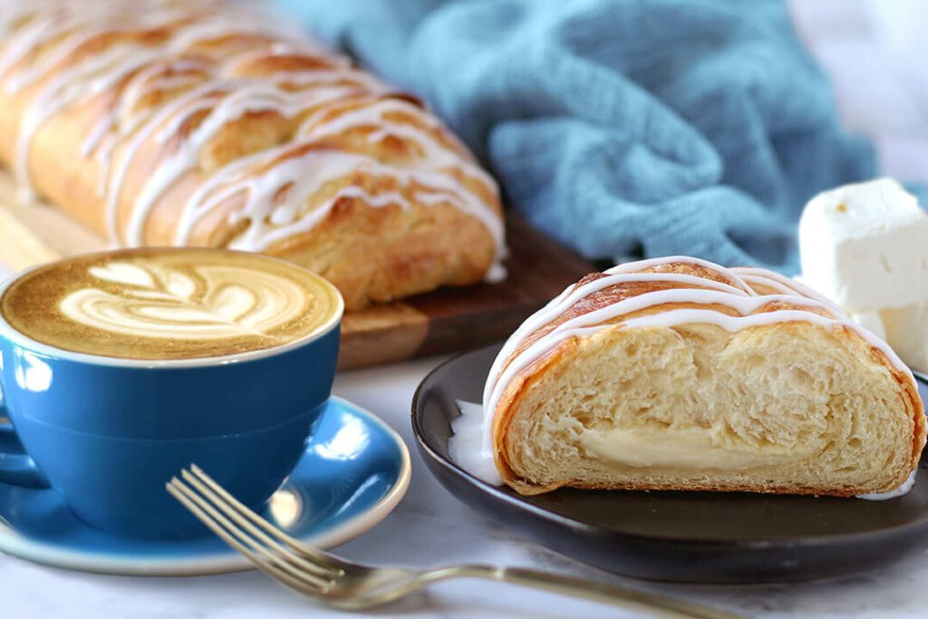 Whole Cream Cheese pastry on cutting board. Slice of pastry on a plate with cream cheese cubes next to it as garnish. Mug with coffee to the side.