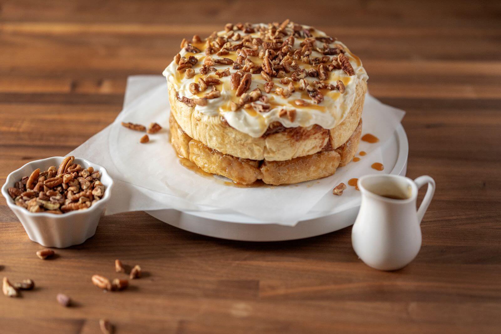 Double-Stacked Pastry Roll Cake on cutting board with bowl of pecans and container of caramel sauce.