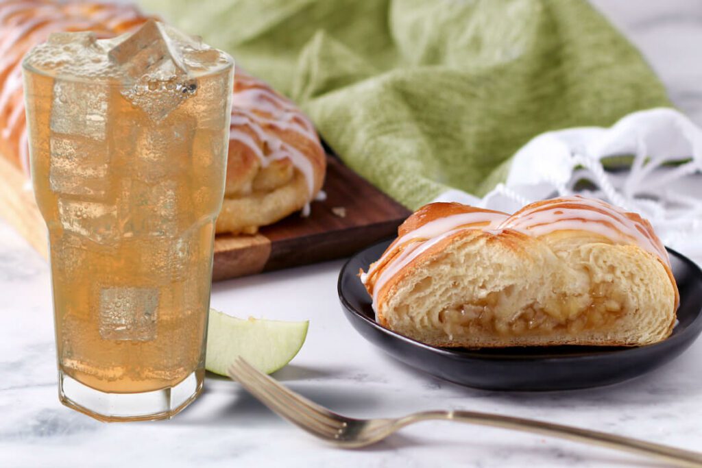 Apple Braided Pastry next to a glass of Apple Cider Spritzer. A full pastry is in the back on a serving platter with a slice on an individual plate. Green cloth napkin is in the background.