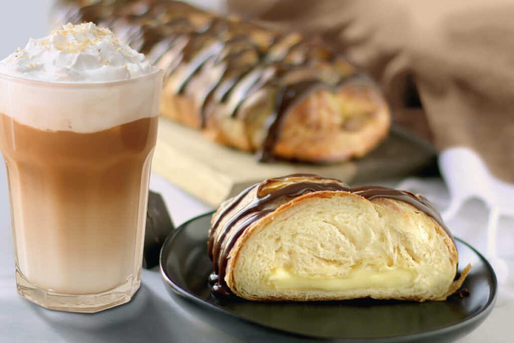 Bavarian Crème Pastry next to a hazelnut latte. A full pastry is in the back on a serving platter with a slice on an individual plate. Brown cloth napkin is in the background.