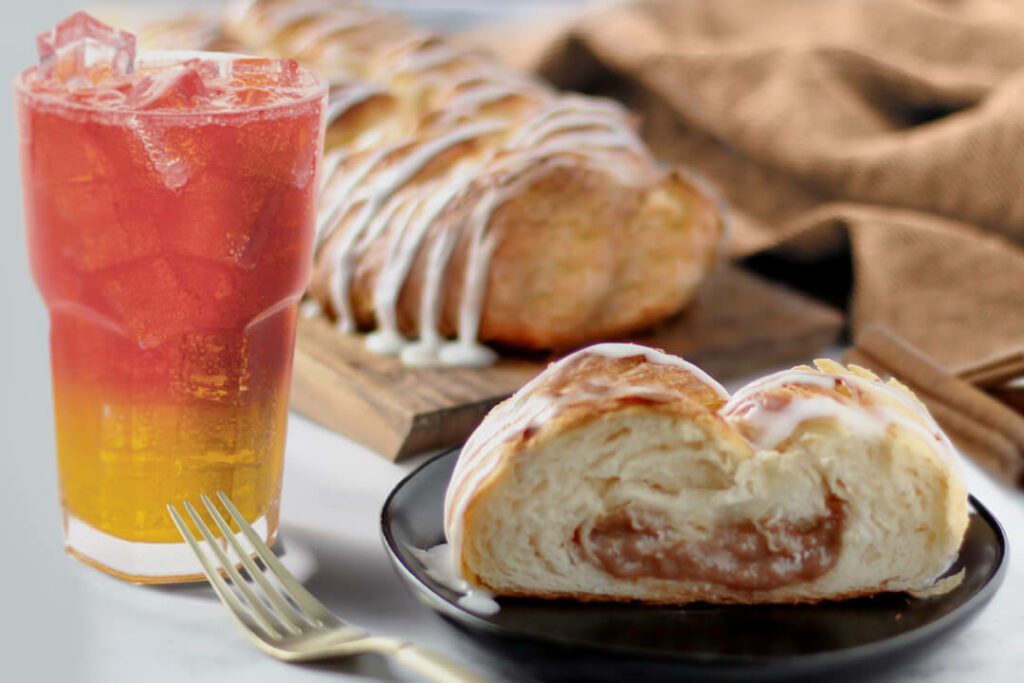 Cinnamon Braided Pastry next to a glass of Cranberry Orange Spritzer. A full pastry is in the back on a serving platter with a slice on an individual plate. Brown cloth napkin is in the background.
