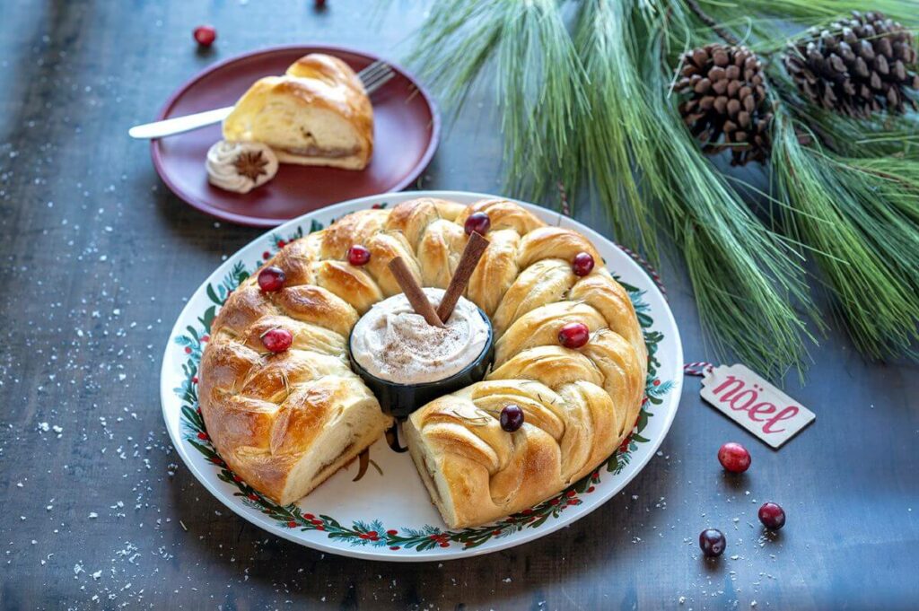 Cinnamon Braided Pastry Ring festively decorated for the Christmas Holiday. A Cinnamon Cream Cheese Dip is in the center of the ring. There are cranberries on the ring. A slice of the pastry is on a plate in the background.