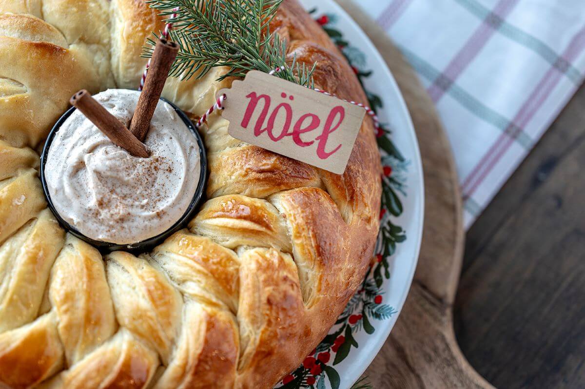 Cinnamon Braided Pastry Ring festively decorated for the Christmas Holiday. A Cinnamon Cream Cheese Dip is in the center of the ring. "Noel" sign is on the braid.