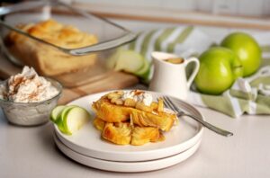 Caramel Apple French Toast slices on a plate. On top is whipped cream and baked apple slices. In the back is a pan with the rest of the Apple pastry loaf. There's also a dish with whipped topping, one with the homemade syrup, and green apples.