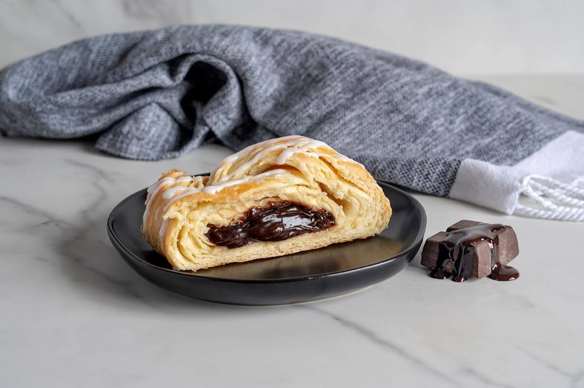 Slice of the Chocolate Braided Pastry on a black plate next to a square of chocolate with chocolate syrup dripping on it. There's a gray napkin in the background.