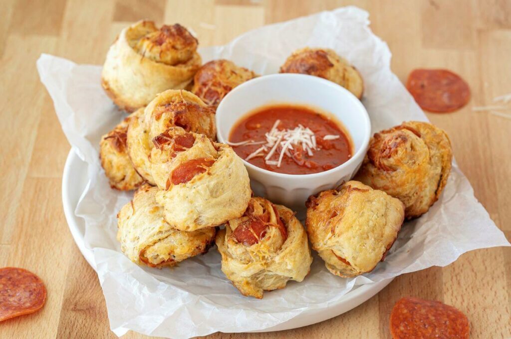 A plate full of Four Cheese Pepperoni Pinwheels with a bowl of marinara sauce in the center. The plate is on a wood background which has pepperonis and cheese.