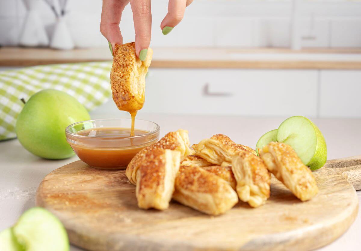 Apple pastry fries on a wooden serving board. There's a bowl of dipping sauce next to the fries, and a hand is dipping one of the fries into the sauce.