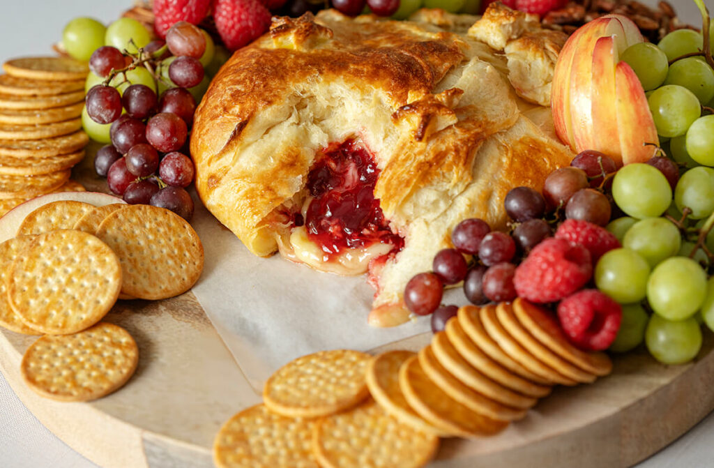 Raspberry Brie Pastry on a charcuterie board surrounded by grapes, raspberries, apple slides, and crackers. Part of the dish has been cut out, revealing the filling.