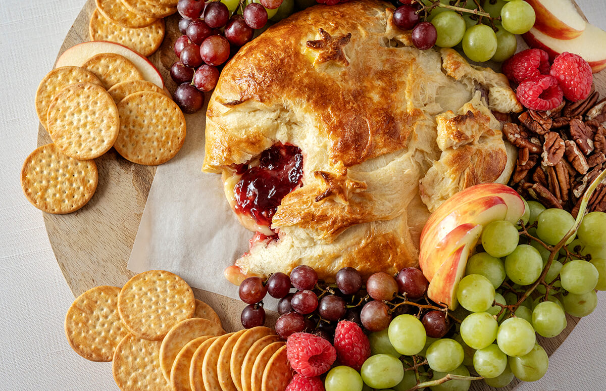 Raspberry Brie Pastry on a charcuterie board surrounded by grapes, raspberries, apple slides, and crackers. Part of the dish has been cut out, revealing the filling.