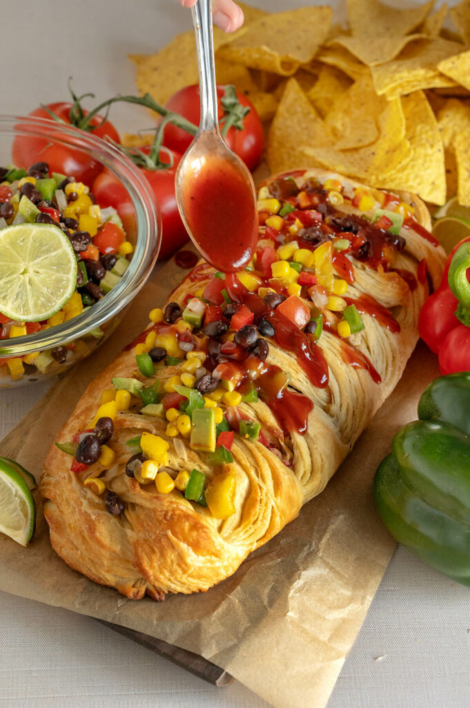 Image of someone spooning raspberry BBQ sauce overtop the Raspberry Cowboy BBQ Pastry. The pastry is surrounded by a bowl of cowboy caviar, tortilla chips, and vegetables.