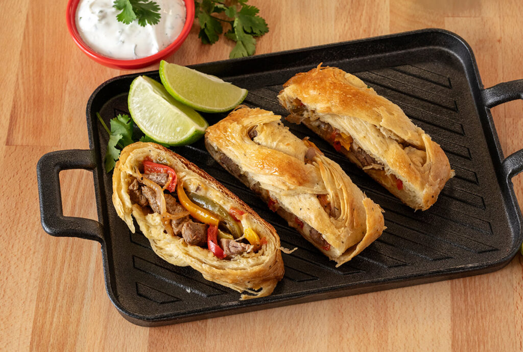 Three slices of Fajita Pastry on a cast iron serving tray. One of the slices is turned up showing the filling. There are also some lime slices and a bowl of cilantro lime crème.