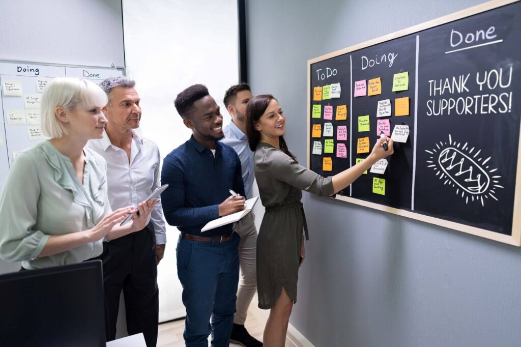 Group of fundraising supporters getting a behind the scenes tour of an organization they sponsor - woman showing them what it is they're working on at a blackboard that has three columns with sticky notes on it. One column has "Thank you supporters!" written on it with a drawing of a pastry.