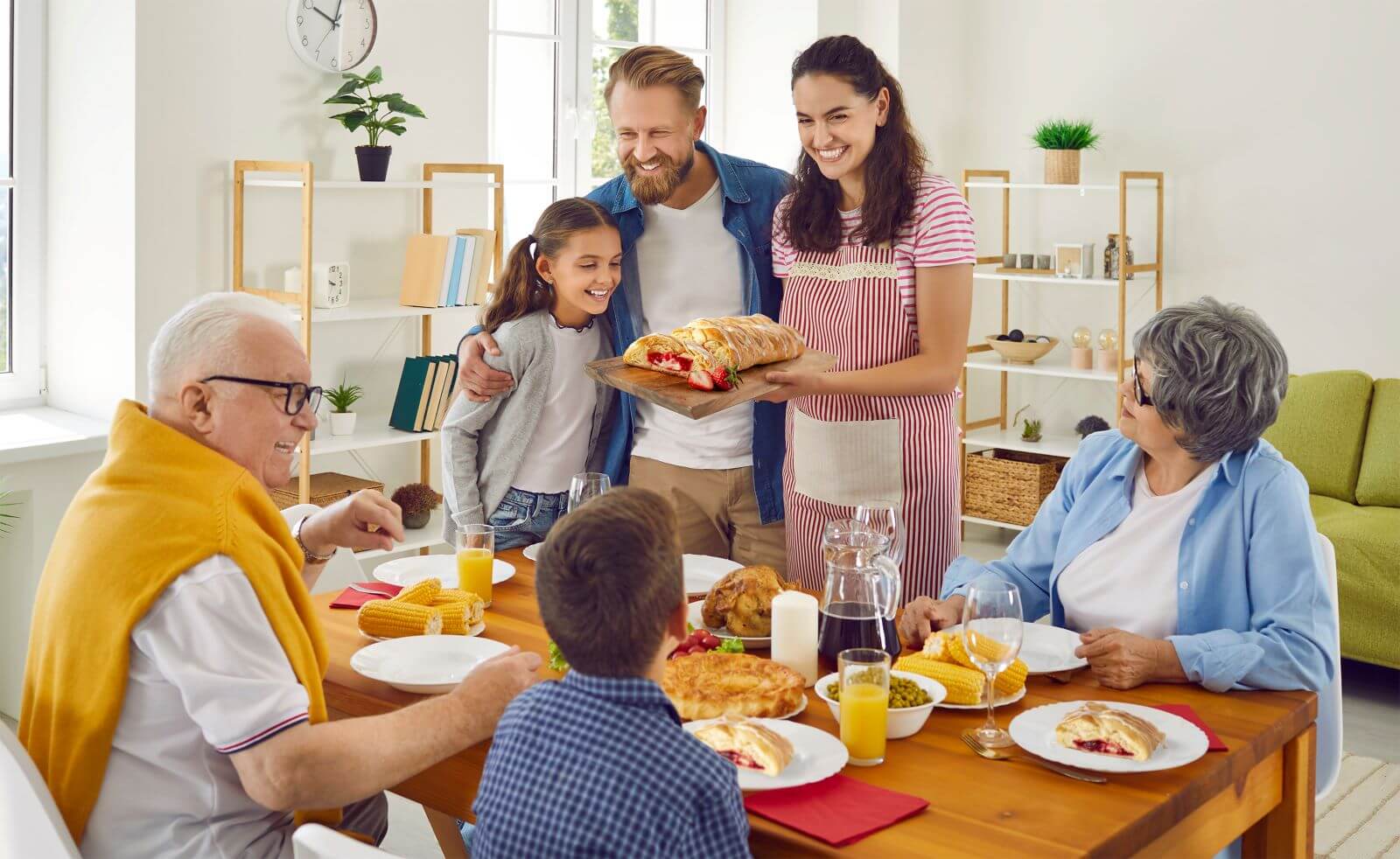 A family gathered around the dining table. Grandpa, Grandma, and the son are sitting down. The mom, dad, and daughter are standing. The mom is holding a Butter Braid Pastry on a serving platter.