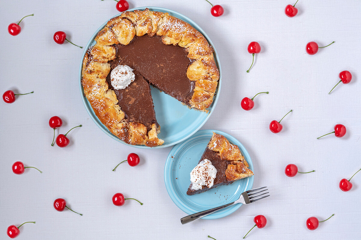 A Black Forest Galette Pastry on a blue serving plate with one slice taken out of it. That slice is on a small blue plate next to the serving plate. Cherries surround the dessert.