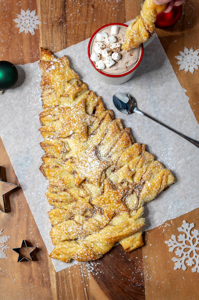 Christmas Tree pastry on large wood cutting board. It's on a piece of parchment paper. Around it are Christmas decorations, snowflakes, and star shaped cookie cutters. A mug of Hot Chocolate Dip is next to it on the parchment paper. A hand is dipping a piece of the pastry in the Hot Chocolate Dip.