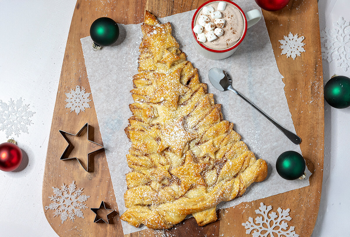 Christmas Tree pastry on large wood cutting board. It's on a piece of parchment paper. Around it are Christmas decorations, snowflakes, and star shaped cookie cutters. A mug of Hot Chocolate Dip is next to it on the parchment paper.