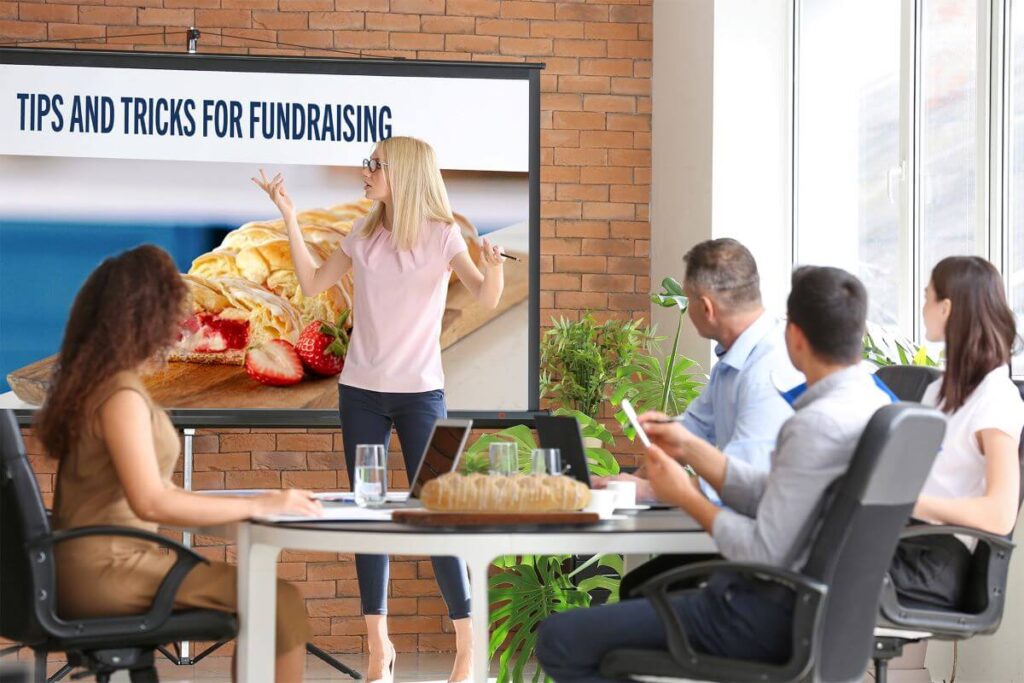 A fundraising representative kicking off a local group's fundraiser by giving a presentation in a meeting room. There is a Butter Braid® Pastry on the table and a PowerPoint on the screen that says "Tips and Tricks for Fundraising".