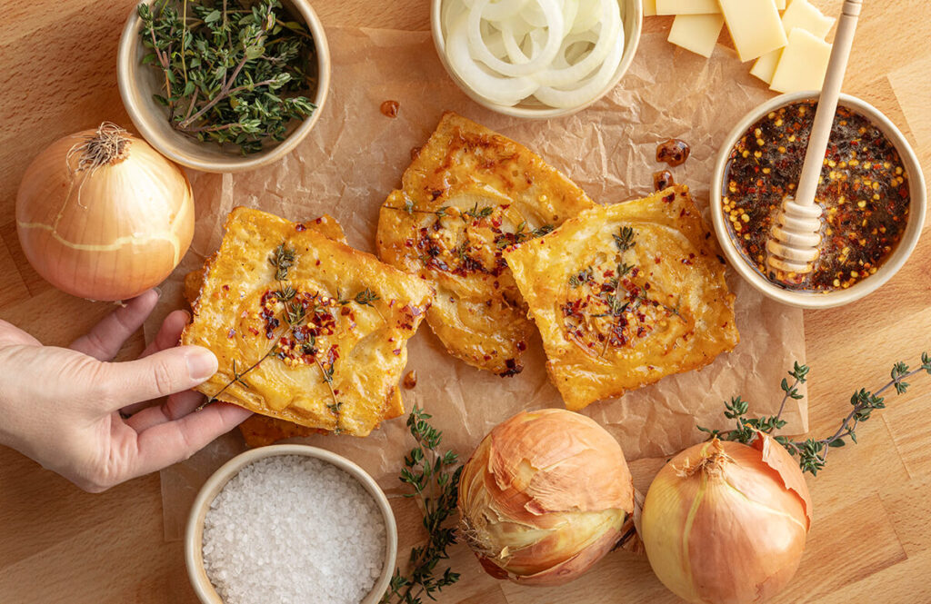 Upside-Down Onion Tarts surrounded by the various ingredients used to make them including onions, thyme leaves, hot honey, gruyere cheese, salt, and onion slices. A hand is holding one of the tarts.