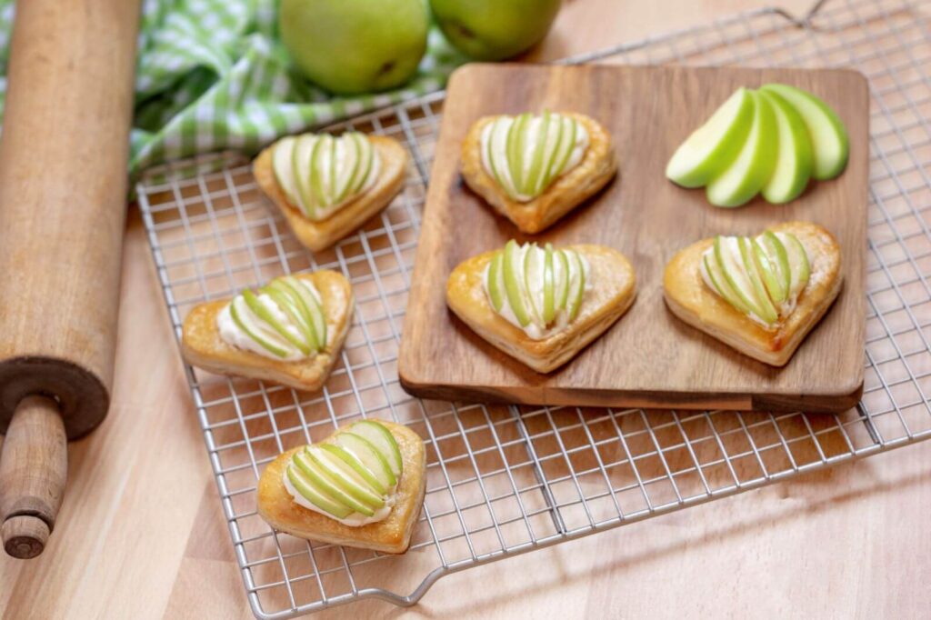 Several Heart-Shaped Apple Pastries on a wire rack. There's also a wood cutting board on the rack with more pastries. There are green apples all around the rack and a wood rolling pin.