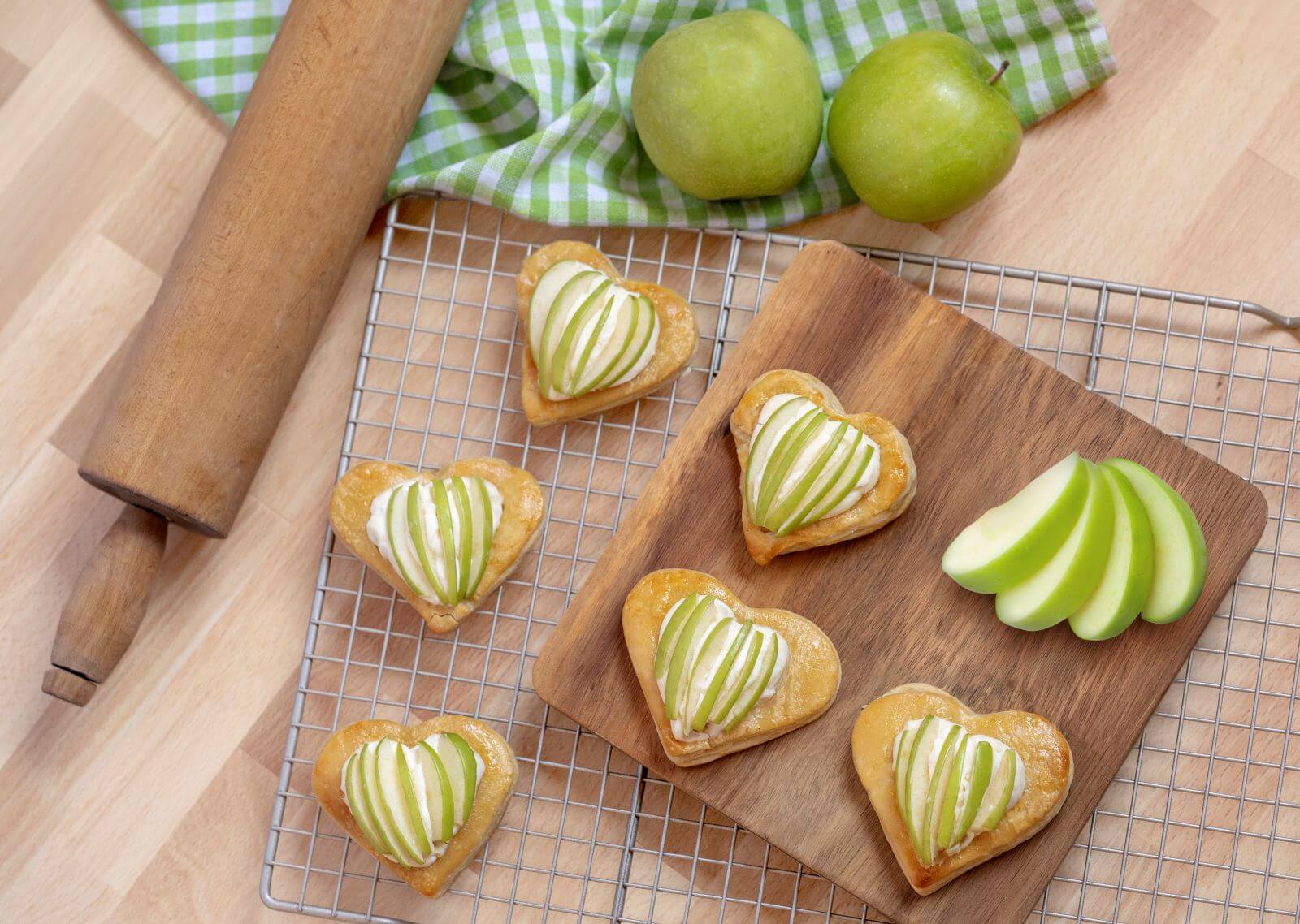 Several Heart-Shaped Apple Pastries on a wire rack. There's also a wood cutting board on the rack with more pastries. There are green apples all around the rack and a wood rolling pin.