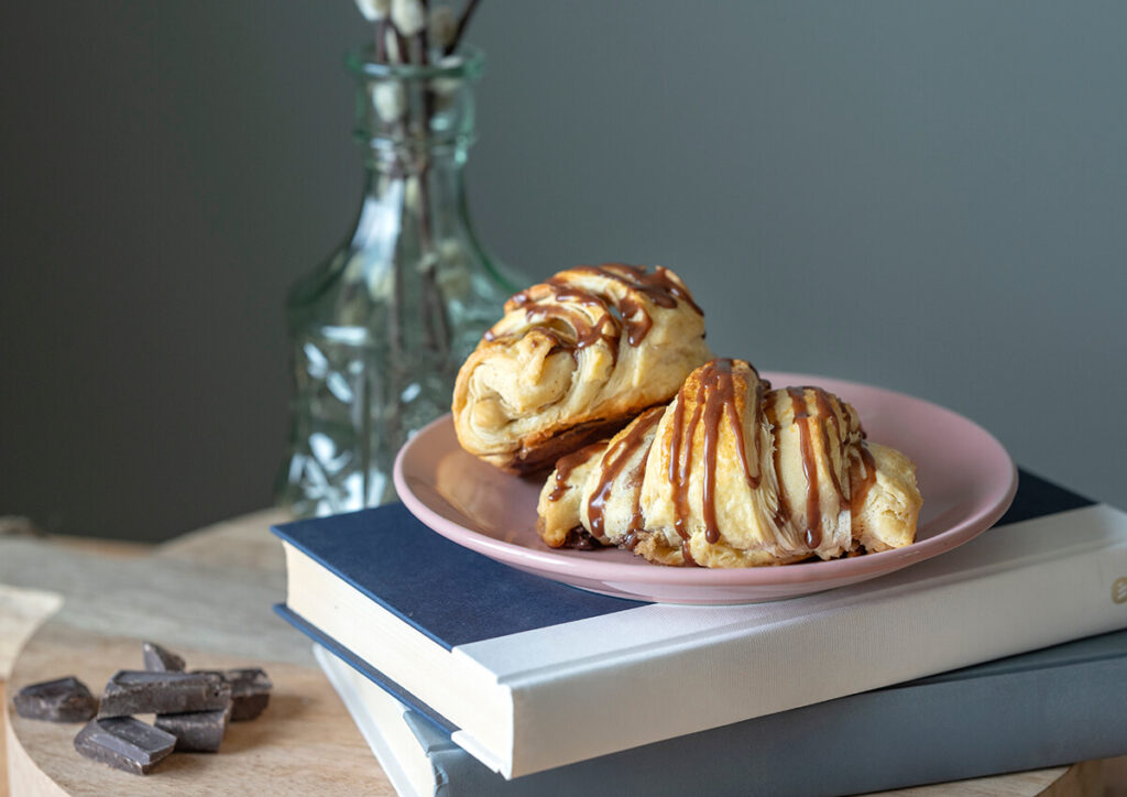 A plate of Bavarian Crème Cookie Croissants stacked on top of two books. In the background is a vase. Next to the books are several chunks of chocolate.