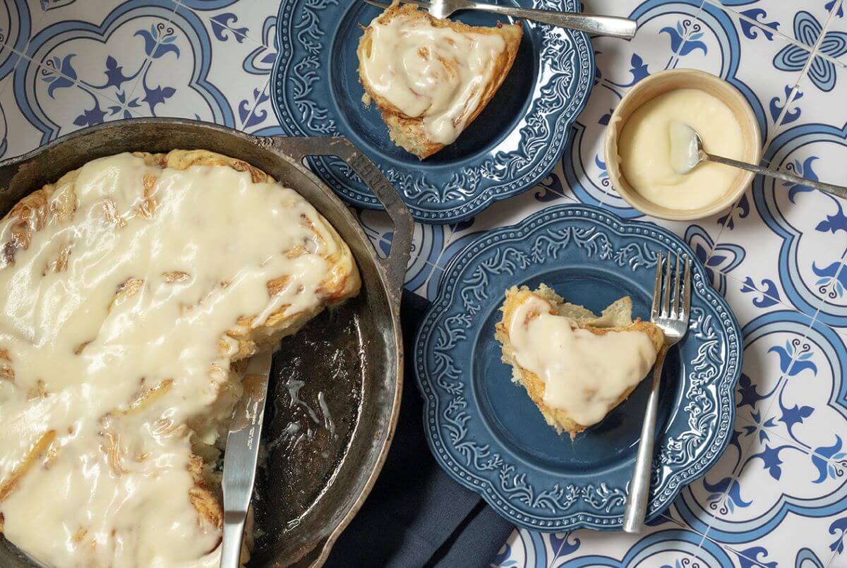 A pan of the viral Cinnamon Pastry Rolls with two taken out of it. Those two are on plates next to the pan. There is a bowl of icing next to those plates.