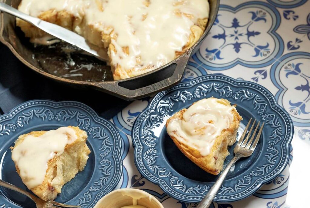 A pan of the viral Cinnamon Pastry Rolls with two taken out of it. Those two are on plates next to the pan. There is a bowl of icing next to those plates.