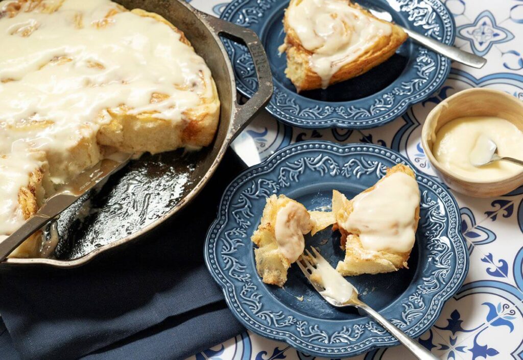 A pan of the viral Cinnamon Pastry Rolls with two taken out of it. Those two are on plates next to the pan. There is a bowl of icing next to those plates.