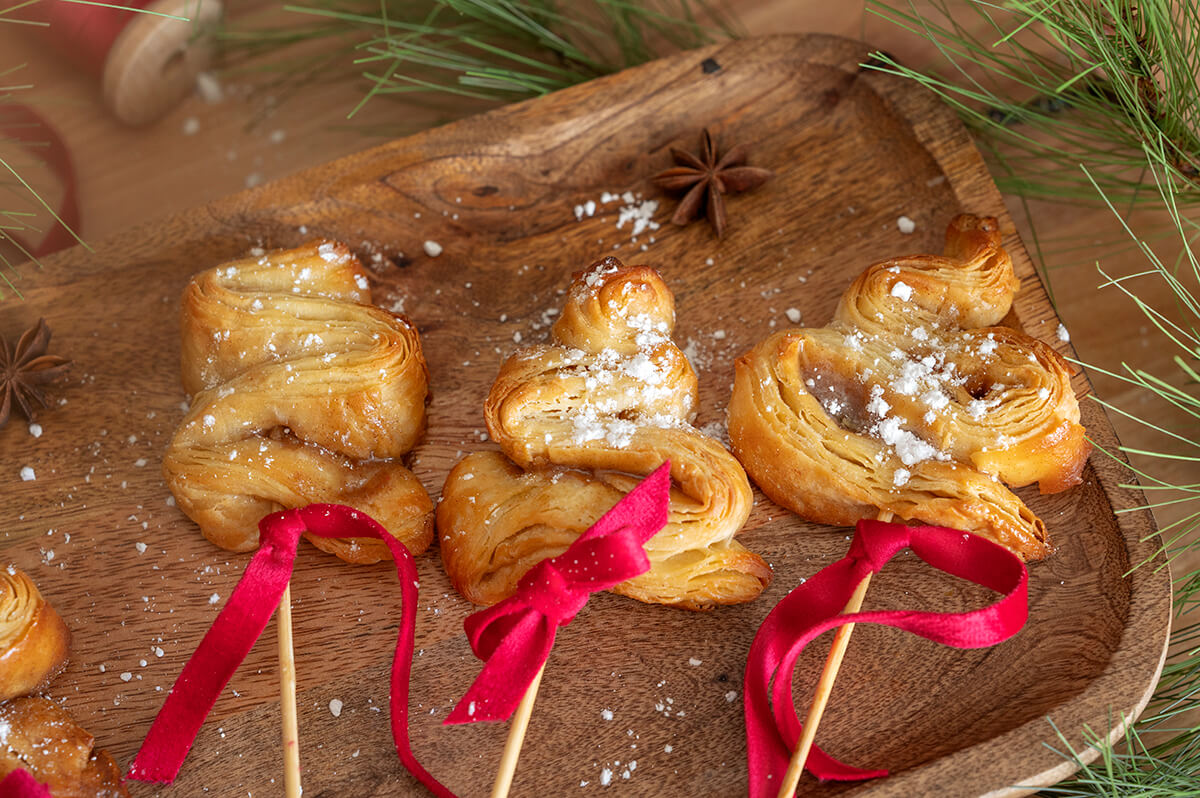 Christmas Tree Pastries on a wooden serving board surrounded by pine needles and star anise. There are red ribbons tied on the skewers.
