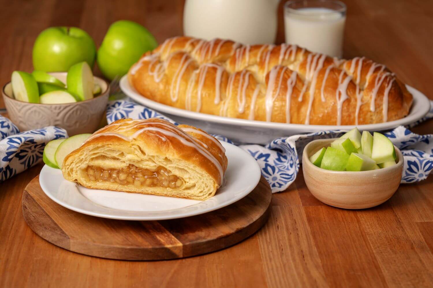 An Apple Butter Braid Pastry on a serving platter. Next to it is a plate with a slice of pastry on it. There are bowls of green apple slices next to glasses of milk.