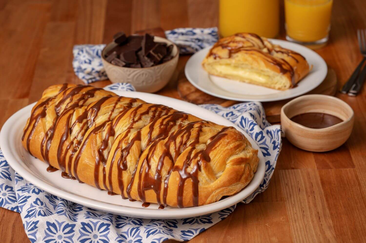 A Bavarian Creme Butter Braid Pastry on a serving platter. Next to it is a plate with a slice of pastry on it. There are bowls of chocolate chunks and chocolate icing next to glasses of orange juice.