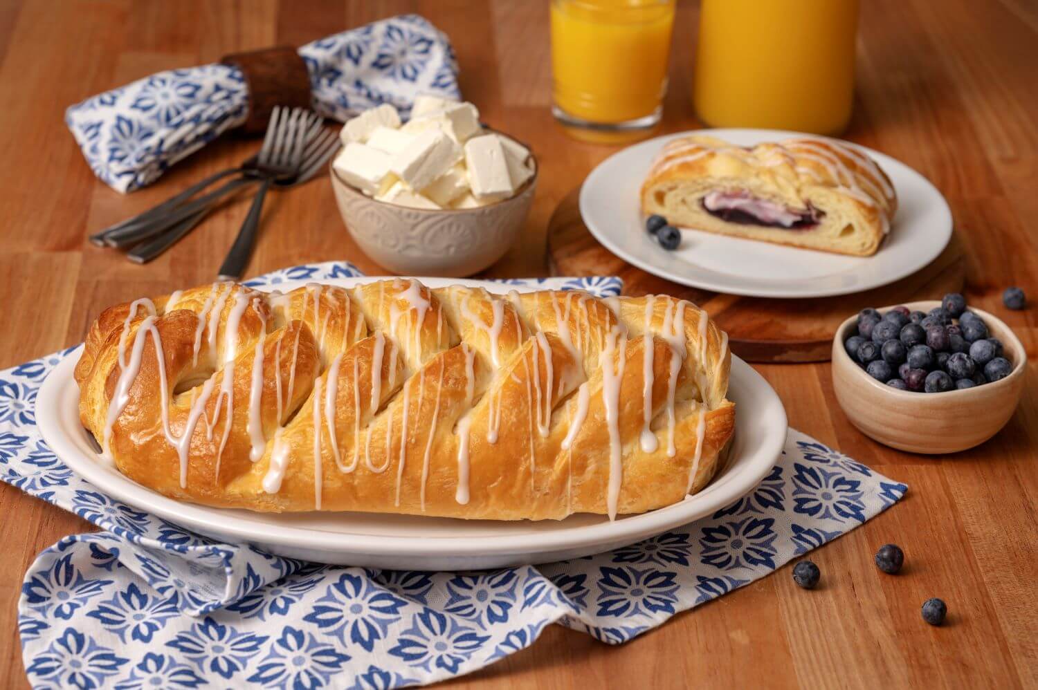 A Blueberry Cream Cheese Butter Braid Pastry on a serving platter. Next to it is a plate with a slice of pastry on it. There are bowls of cream cheese chunks and blueberries next to glasses of orange juice.