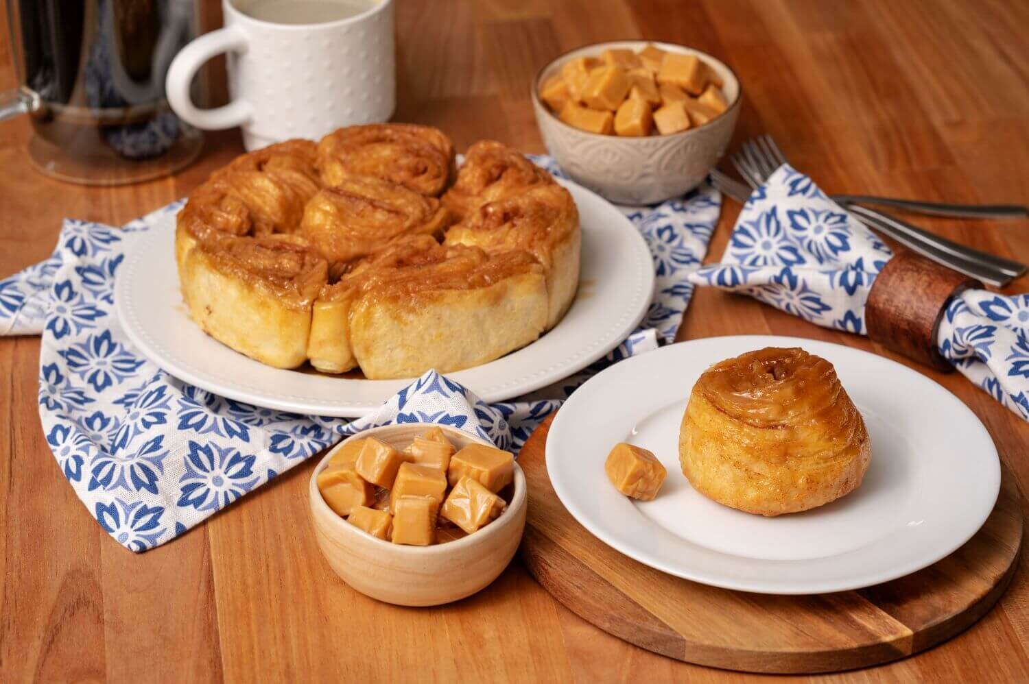 Glazed Caramel Butter Braid Pastry Rolls on a serving platter. Next to it is a plate with a single pastry roll on it. There are bowls of caramel candies next to mugs of coffee.