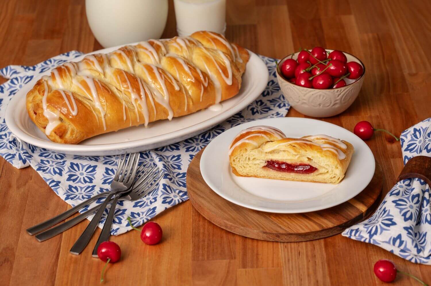 A Cherry Butter Braid Pastry on a serving platter. Next to it is a plate with a slice of pastry on it. There is a bowl of cherries next to glasses of milk.
