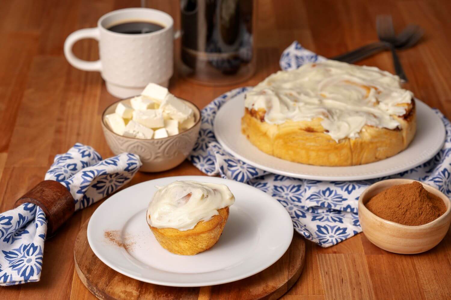 Iced Cinnamon Butter Braid Pastry Rolls on a serving platter. Next to it is a plate with a single pastry roll on it. There are bowls of cream cheese chunks and cinnamon powder next to mugs of coffee.