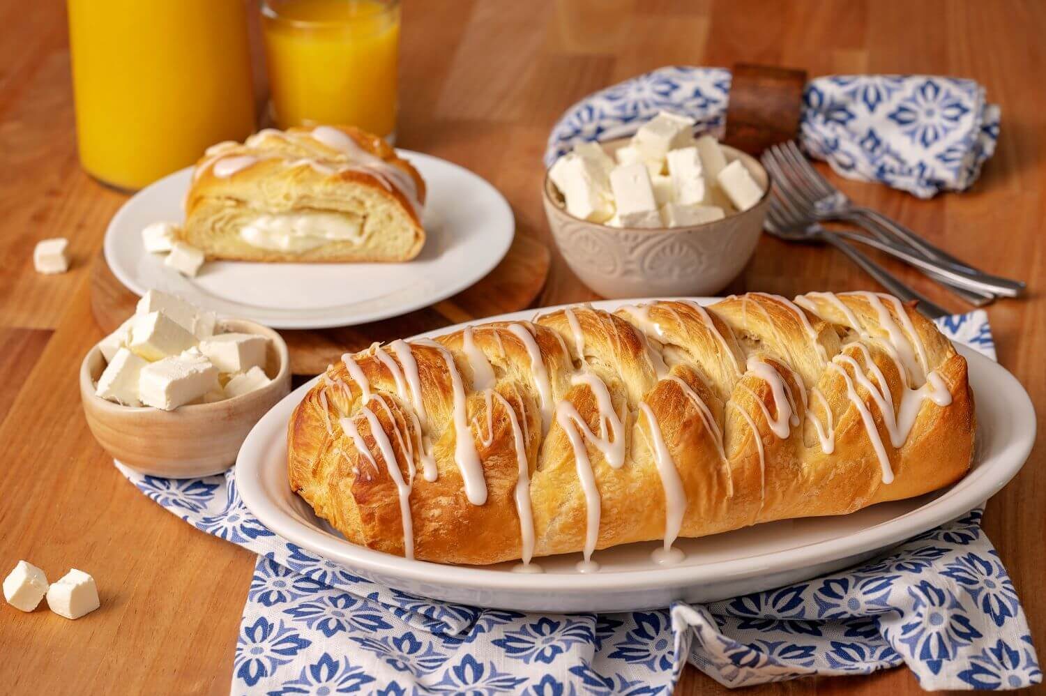 A Cream Cheese Butter Braid Pastry on a serving platter. Next to it is a plate with a slice of pastry on it. There are bowls of cream cheese chunks next to glasses of orange juice.