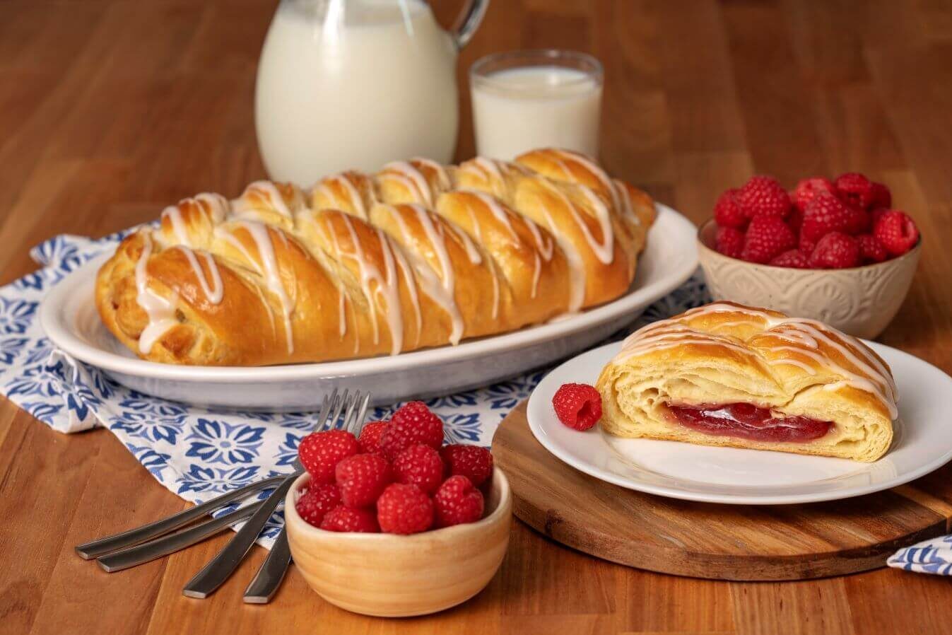 A Raspberry Butter Braid Pastry on a serving platter. Next to it is a plate with a slice of pastry on it. There are bowls of raspberries next to glasses of milk.