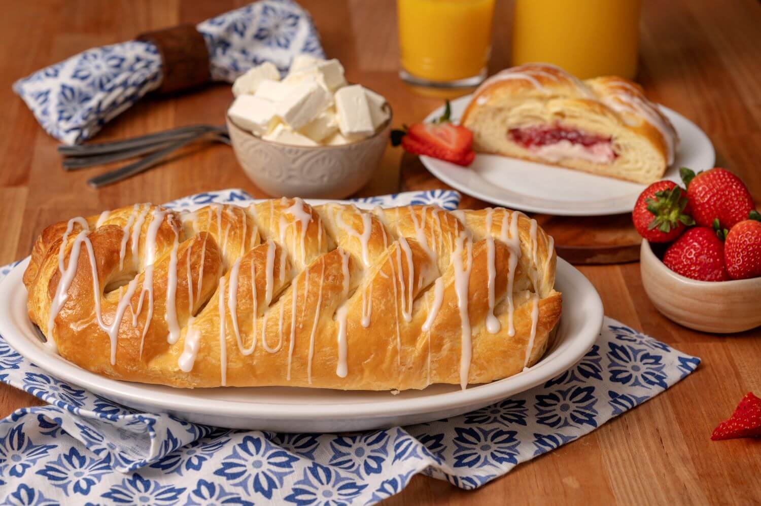 A Strawberry Cream Cheese Butter Braid Pastry on a serving platter. Next to it is a plate with a slice of pastry on it. There are bowls of cream cheese chunks and fresh strawberries in the background next to glasses of orange juice.